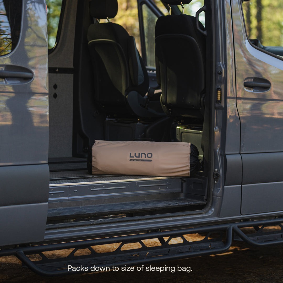 van interior with a LUNO air mattress in the storage bag on the floor, surrounded by trees outside.