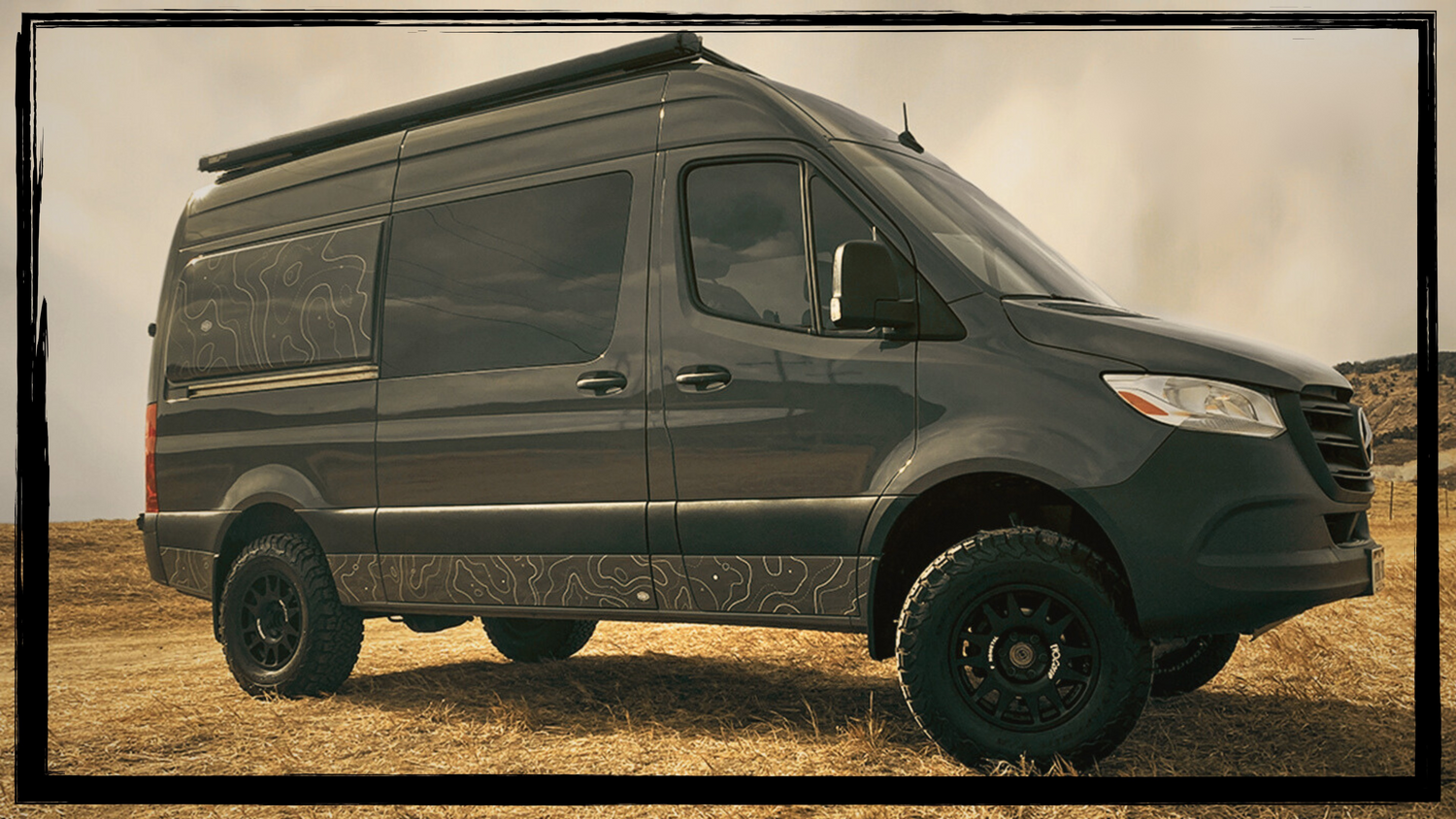 A dark gray custom camper van with off-road tires and a roof rack, parked in a dry grassy field under a hazy sky.